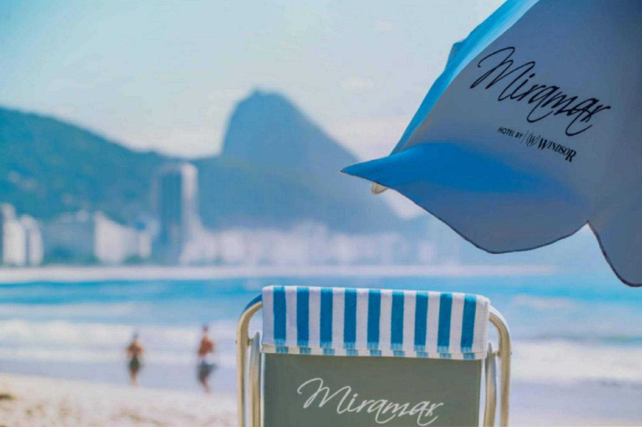فندق ميرامار باي وندسور ريو دي جانيرو المظهر الخارجي الصورة The photo features a beach scene with a lounge chair in the foreground. The chair has a striped blue and white design and displays the name "Miramar." In the background, there are blurred figures walking along the beach, with a hazy view of mountains